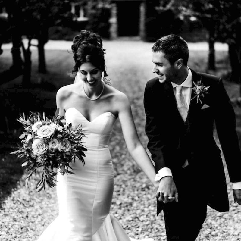 A bride and groom walk hand in hand on a gravel path, wearing formal wedding attire. The bride carries a bouquet and both are smiling, with trees and blurred background—capturing the elegant essence of Dodmoor House Wedding Photography.