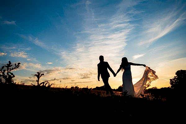 Dodford Manor Wedding Photographer captures the heartfelt moment of a bride and groom holding hands in a picturesque field at sunset.