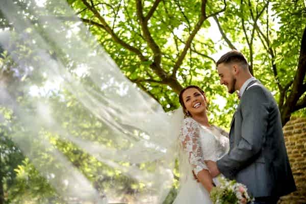 A bride and groom standing under a tree with their veil blowing in the wind, captured by Dodford Manor Wedding Photographer.