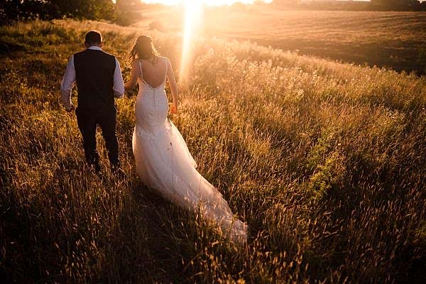 A Dodford Manor wedding photographer captures a beautiful moment as the bride and groom walk through a field at sunset.