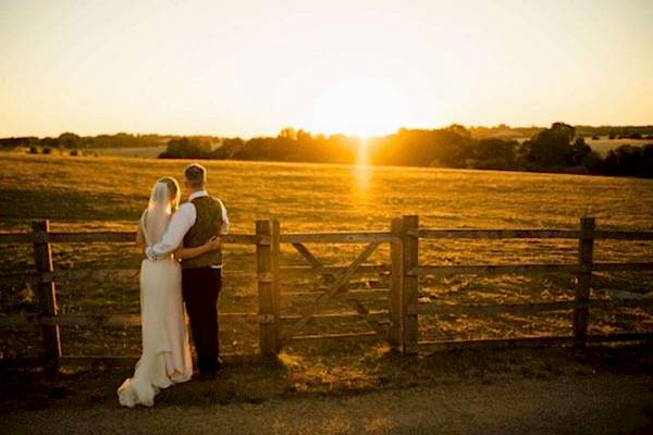 A Dodford Manor Wedding Photographer captures a bride and groom standing in front of a fence at sunset.