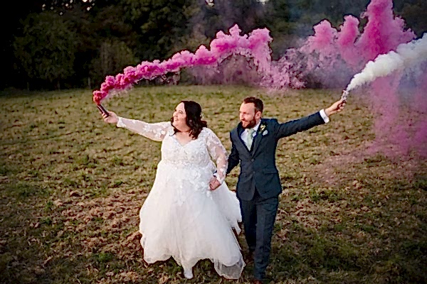 A Dodford Manor Wedding Photographer captures a stunning moment of a bride and groom holding pink smoke bombs in a field.