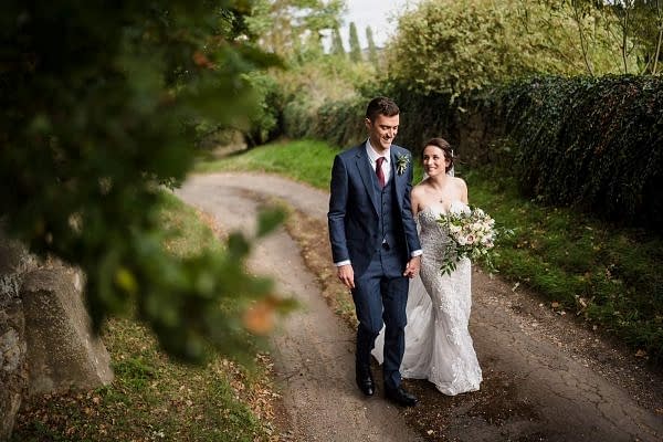 A Dodford Manor Wedding Photographer captures a beautiful moment of a bride and groom walking down a dirt path.