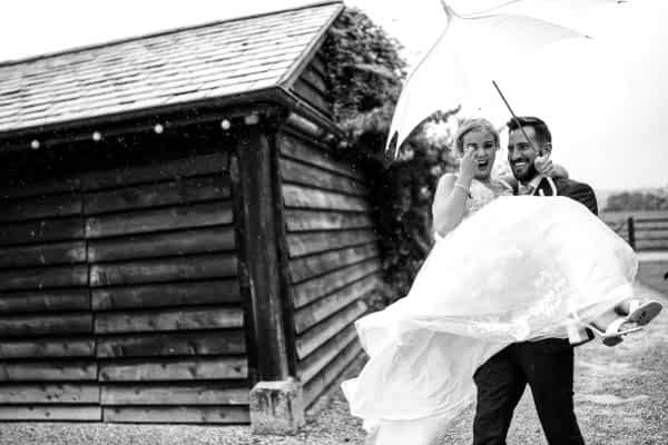 Dodford Manor Wedding Photographer captures a beautiful moment of a bride and groom embracing under an umbrella in the rain.