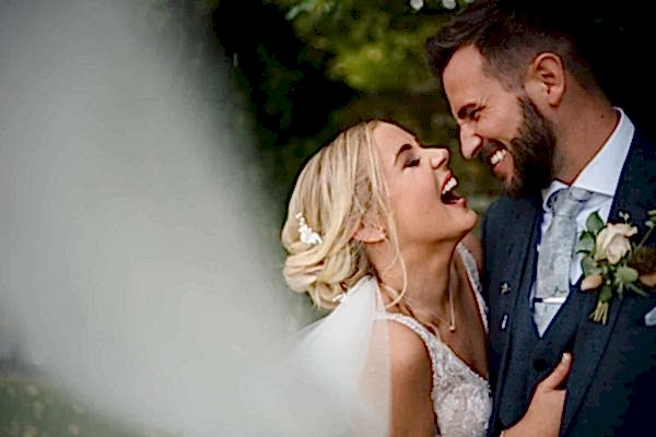 A bride and groom laughing in front of a veil captured by a Dodford Manor Wedding Photographer.
