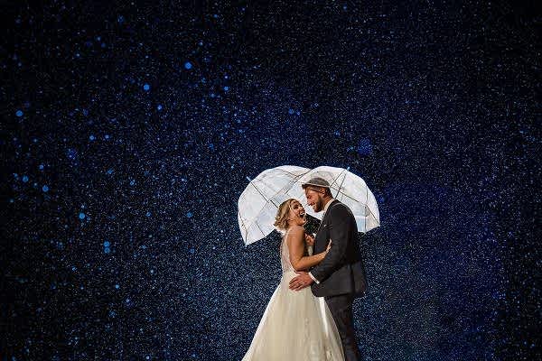 A bride and groom standing under an umbrella in the rain, captured by a Dodford Manor Wedding Photographer.