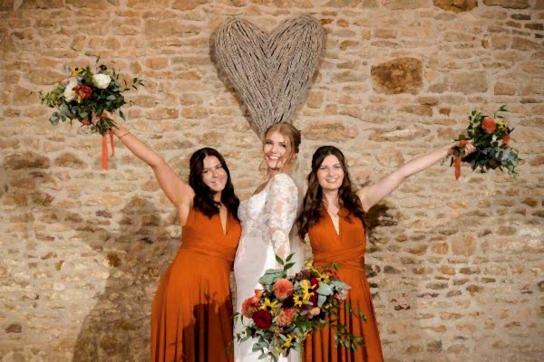 Three bridesmaids holding bouquets at a Dodford Manor wedding, posing elegantly in front of a stone wall.