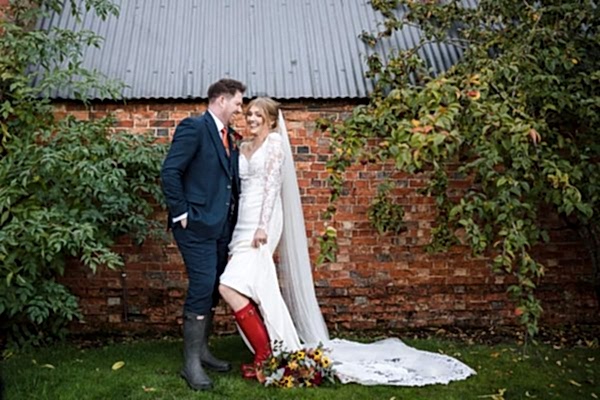 A Dodford Manor Wedding Photographer captures a bride and groom posing in front of a brick wall.