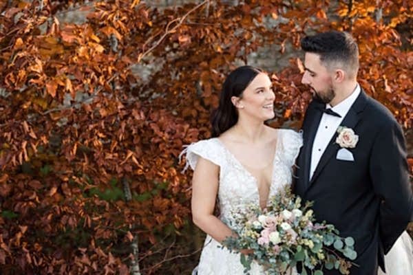 A Dodford Manor Wedding Photographer captures a bride and groom standing in front of a tree in autumn.