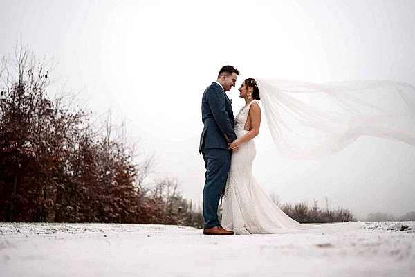 A bride and groom, captured by a Dodford Manor Wedding Photographer, standing in the snow with their veil blowing in the wind.