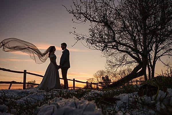 A bride and groom standing in the snow with their veil blowing in the wind captured by a Dodford Manor Wedding Photographer.