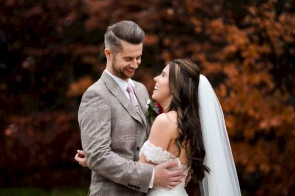A bride and groom embracing in front of a tree, captured beautifully by a Dodford Manor Wedding Photographer.