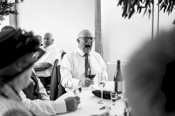 A Dodford Manor Wedding Photographer captures a black and white photo of a man laughing at a table.