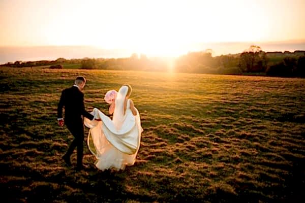 A bride and groom walking in a field at sunset captured by a Dodford Manor Wedding Photographer.