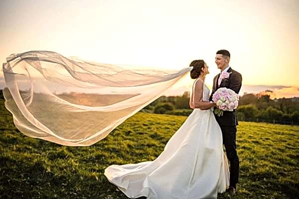 Dodford Manor Wedding Photographer captures a mesmerizing moment of a bride and groom standing in a field with their veil blowing in the wind.