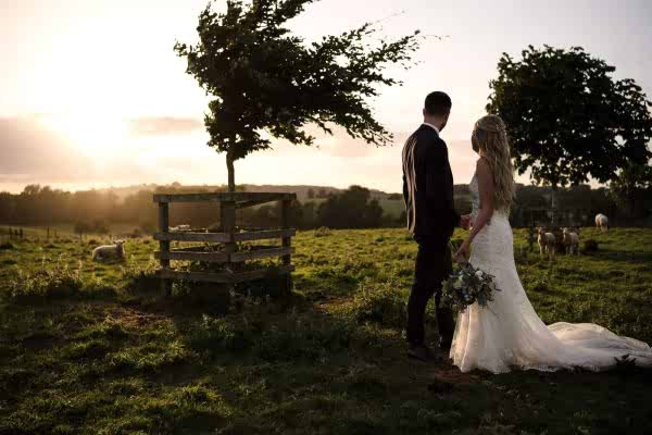 A Dodford Manor Wedding Photographer captures a bride and groom standing in a field at sunset.