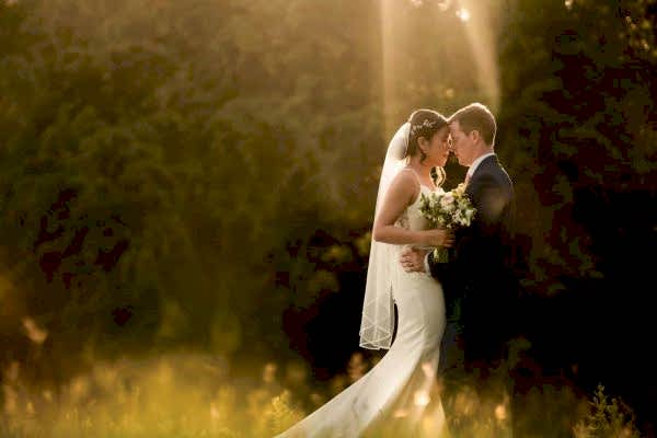 Dodford Manor Wedding Photographer captures a breathtaking moment of a bride and groom kissing in a field at sunset.