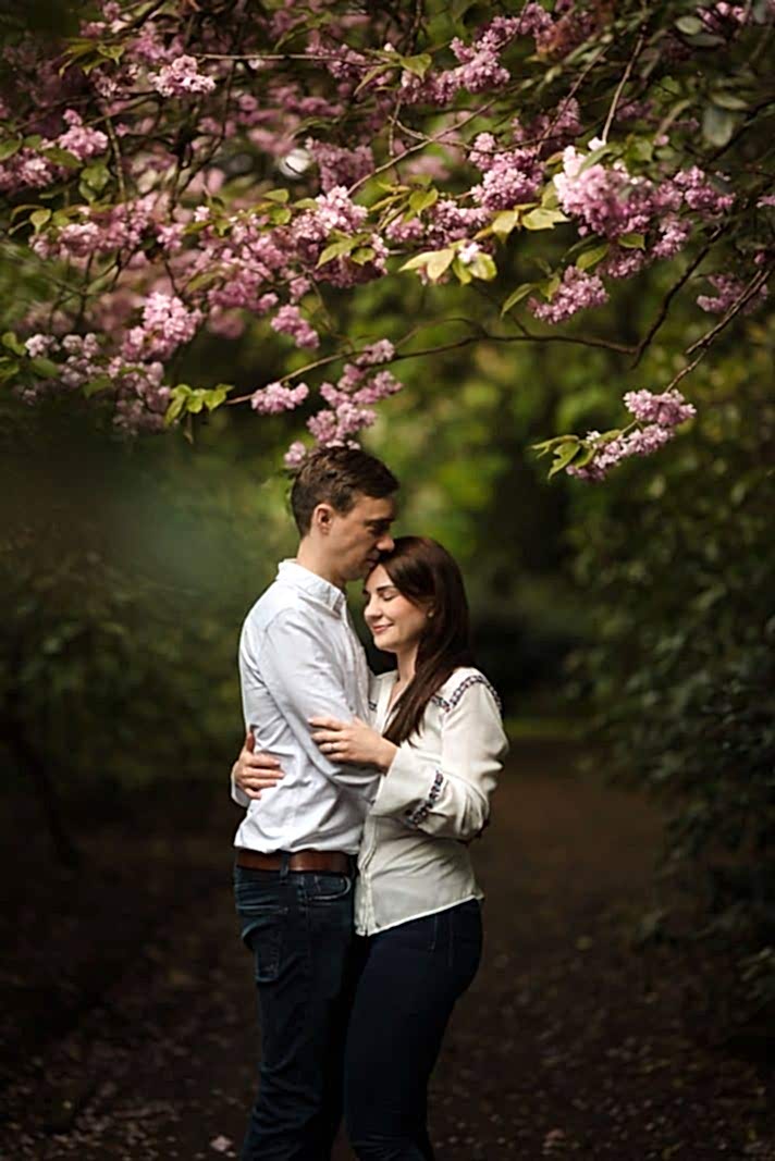 A couple embraces under blooming pink flowers on a tree branch in a lush, green garden, perfectly captured by an engagement photographer in Northamptonshire.