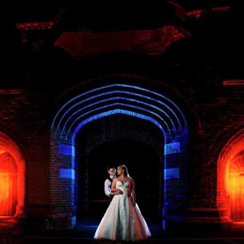 A bride and groom stand embraced under a gothic archway with dramatic red and blue lighting illuminating the scene, capturing a timeless moment of Holmewood Hall Wedding Photography.