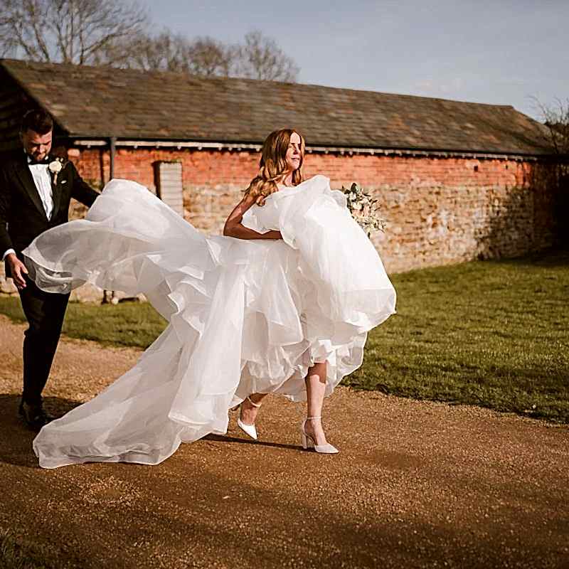 A bride in a white gown walks on a path in front of a rustic building, holding her dress, while a groom in a tuxedo follows closely, assisting with the gown's train.