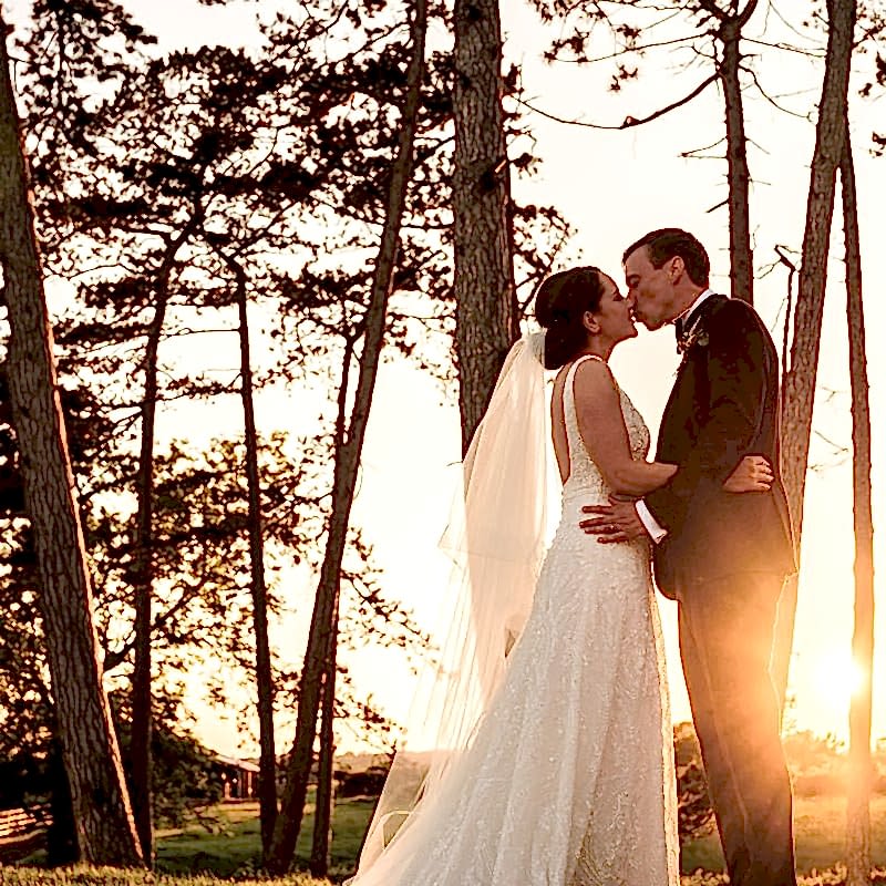 A couple in wedding attire shares a kiss in front of tall trees with sunlight streaming through, creating an ethereal glow in a natural outdoor setting, beautifully captured by a Leicestershire wedding photographer.