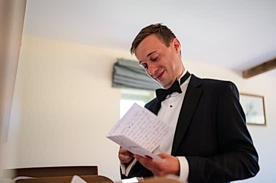 A person in a tuxedo is smiling and reading a card in a room with bright lighting, showcasing the benefits of two wedding photographers capturing every joyful moment from different angles.