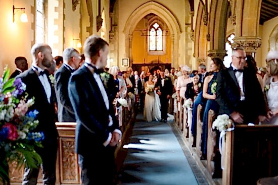 The bride and groom gracefully walk down the aisle of a Cambridgeshire church, surrounded by standing guests in formal attire, beautifully captured by the benefits of two wedding photographers ensuring every cherished moment is immortalized.