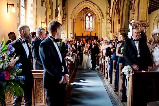 A bride and groom walk down the aisle in a church filled with seated guests dressed in formal attire, capturing every moment from multiple angles thanks to the benefits of two wedding photographers.