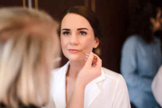 A woman in a white robe is having her makeup applied by another person using a brush. Capturing such intimate moments highlights the benefits of two wedding photographers, ensuring every detail and angle of your special day is perfectly preserved.