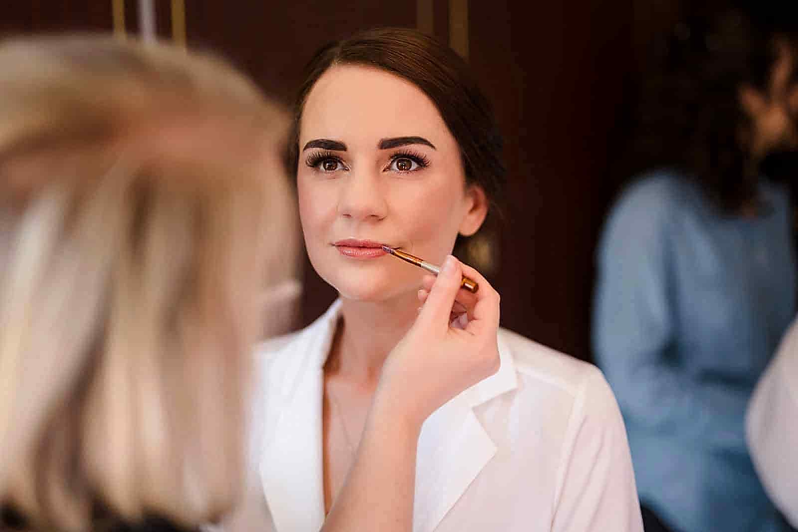 In a well-lit room, a woman in a white top has makeup carefully applied to her lips, while two wedding photographers from Leicestershire capture the enchanting moment.