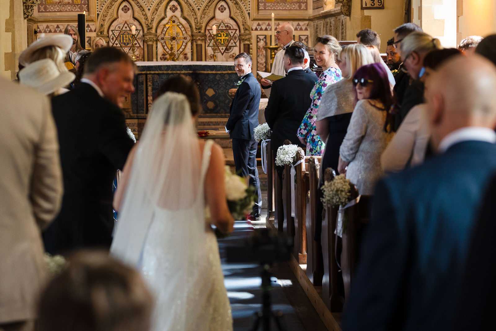 A bride walks down the aisle of a church, surrounded by seated guests, as two wedding photographers based in Leicestershire capture each magical moment. Meanwhile, the groom waits eagerly at the altar.