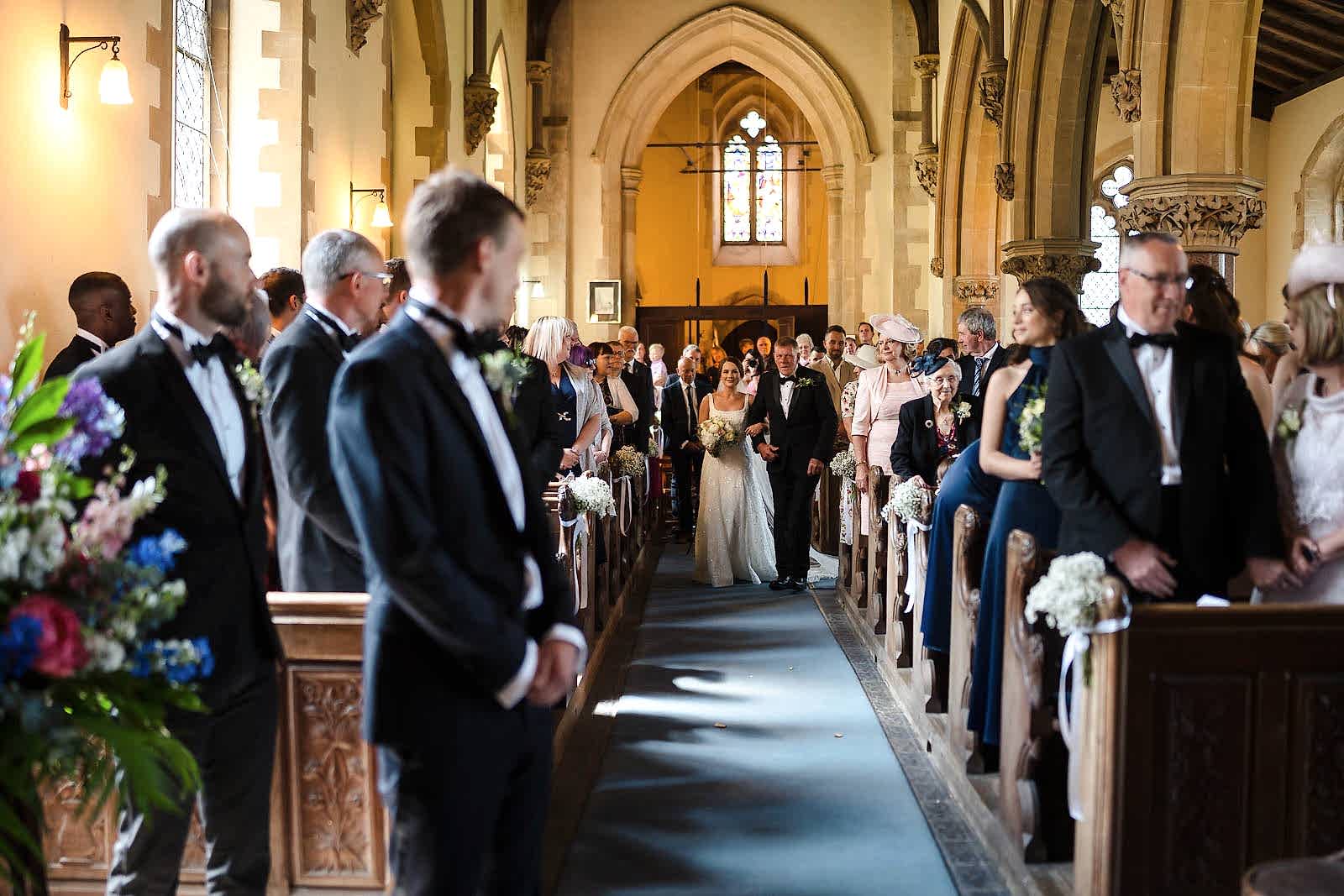 Bride walking down the aisle in a church, guests seated and standing, with two wedding photographers based in Leicestershire capturing every moment alongside two men in suits in the foreground.