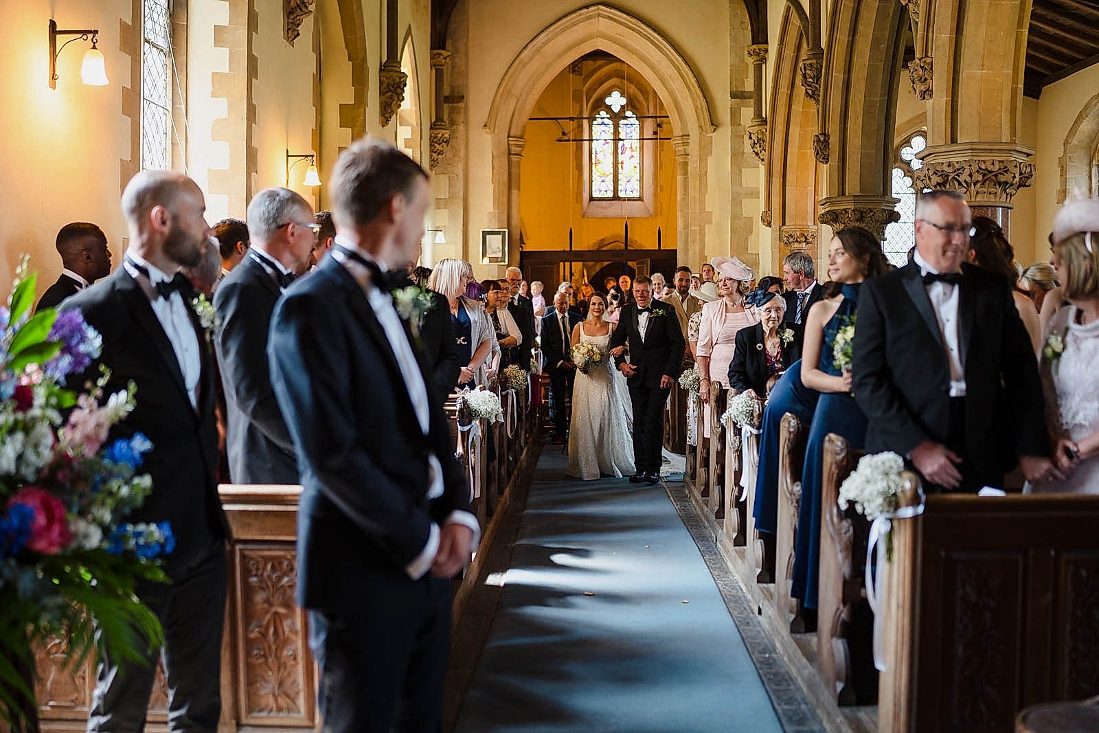 A bride walks down the aisle with her father in a church filled with guests, as two wedding photographers from Bedfordshire discreetly capture each magical moment.