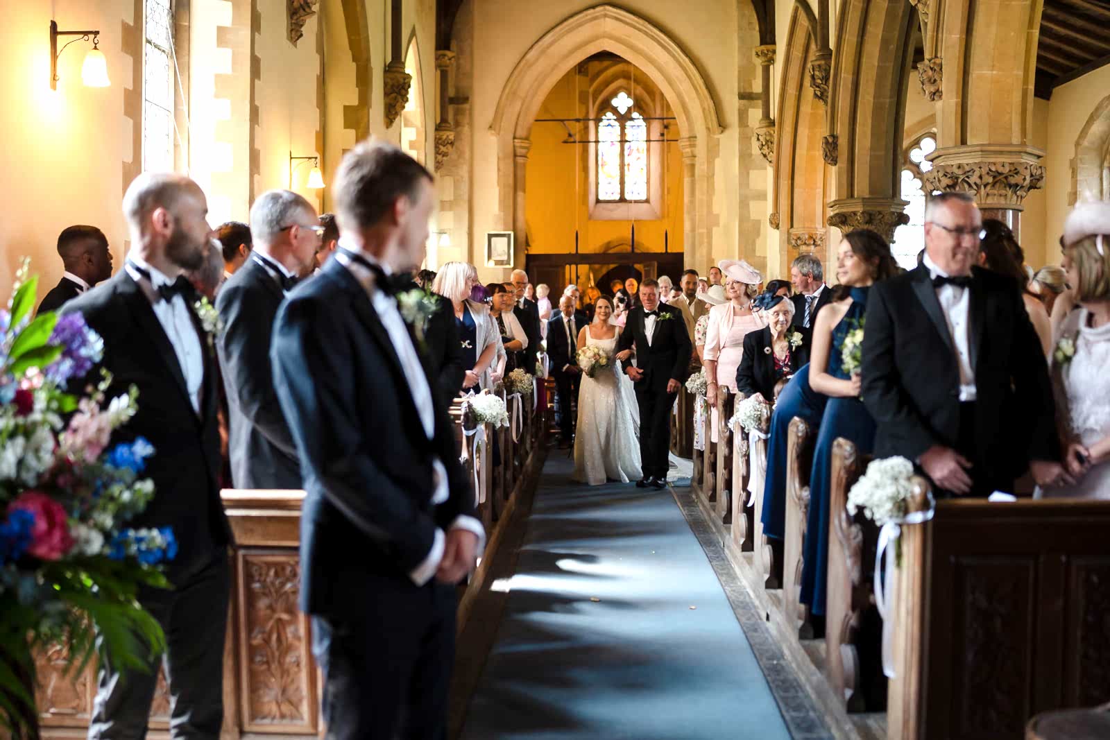 A bride walks down the aisle in a church, accompanied by a man. With two wedding photographers capturing every angle, guests stand on either side, watching the ceremony unfold in all its splendor.