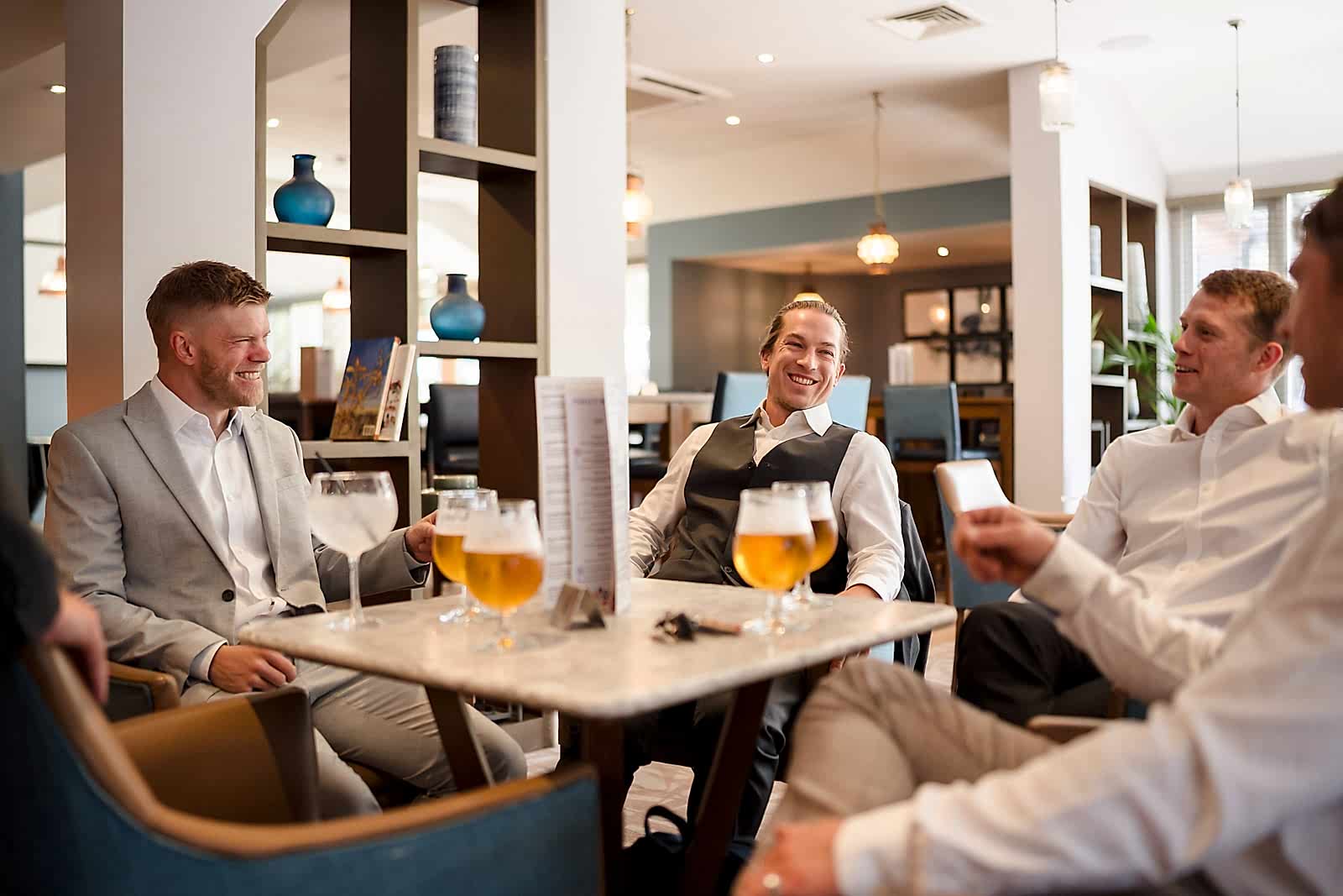 Four men in formal attire sit around a restaurant table with glasses of beer, engaged in conversation. The setting, reminiscent of groom prep photography, offers a relaxed backdrop, adorned with shelves and pendant lights illuminating the camaraderie shared.