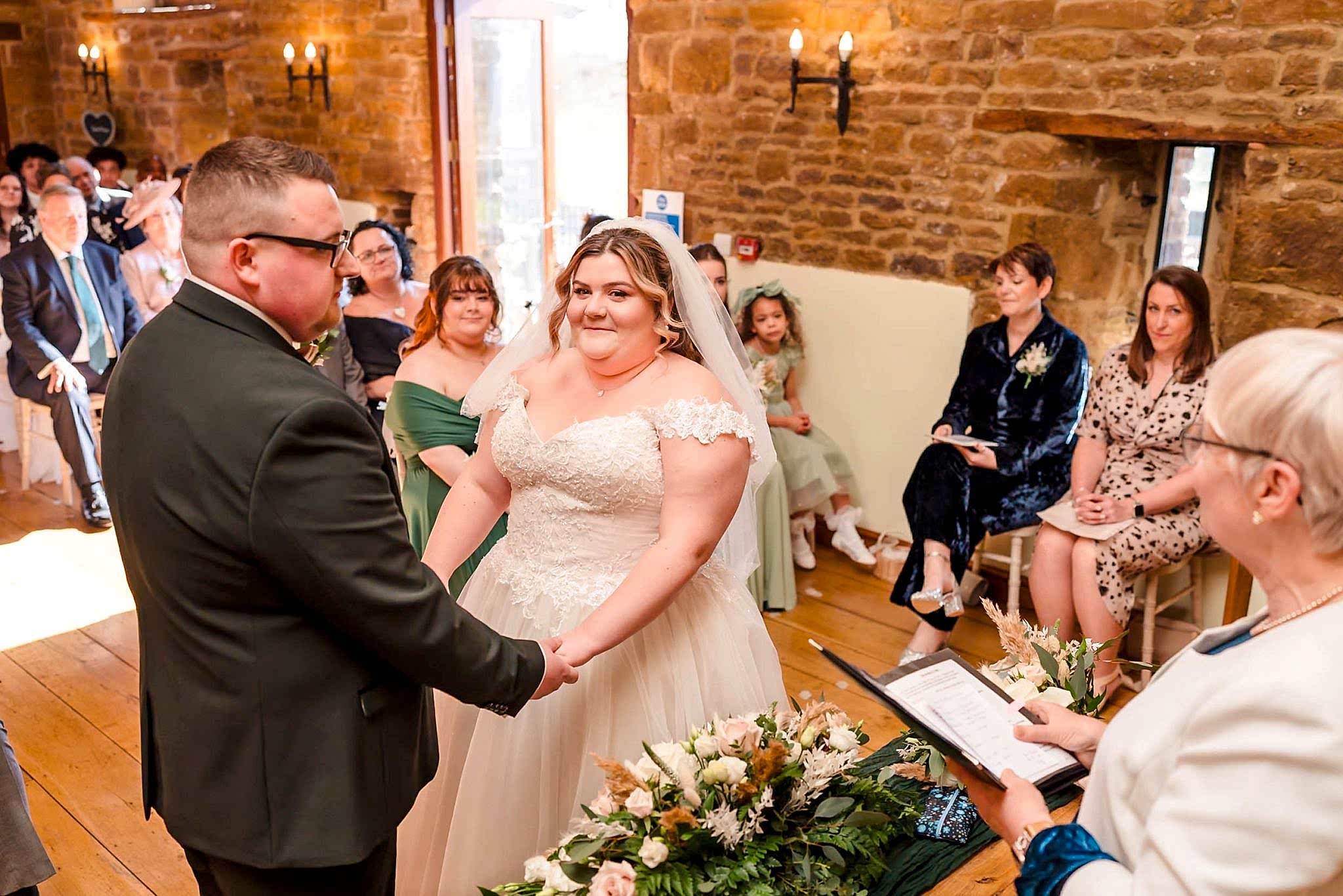 A bride and groom hold hands during a wedding ceremony in a rustic venue. Guests are seated, and an officiant stands nearby with a document. Flowers are arranged on a table in the foreground.